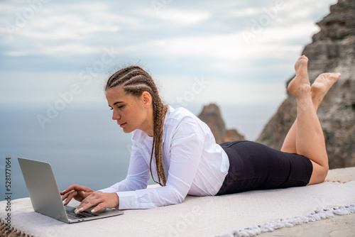 A woman is lying and typing on a laptop keyboard on a terrace with a beautiful sea view. Wearing a white blouse and black skirt. Freelance travel and vacation concept, digital nomad.