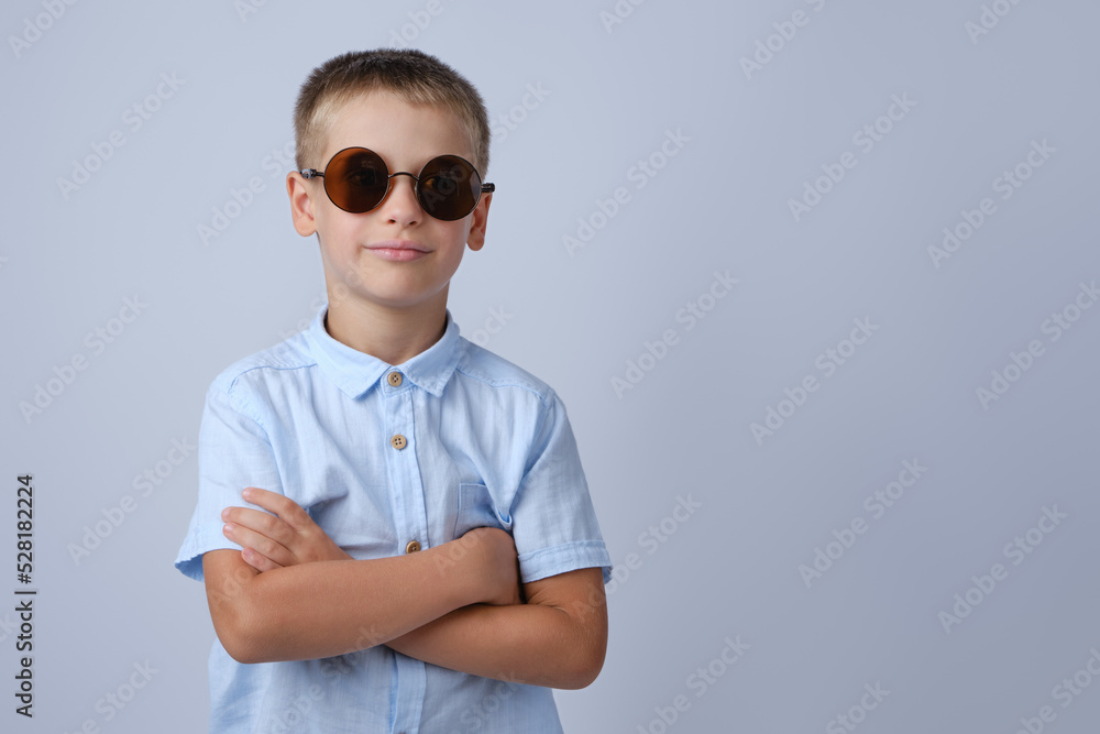 A child in round sunglasses on a gray background with crossed arms. Place for text. A child in round sunglasses on a gray background with crossed arms. Place for text.