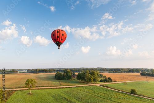 Red hot air balloon with black dots flying over fields and forests on a sunny summer morning. Aerial view, Latvia.