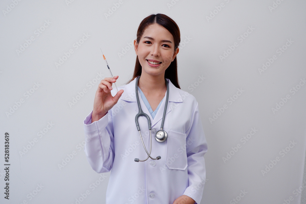 Happy smiling Asian female doctor wearing white medical uniform holding a syringe with a medical stethoscope on gray background medical concept. Happy smiling Asian female doctor wearing white medical uniform holding a syringe with a medical stethoscope on gray background medical concept.