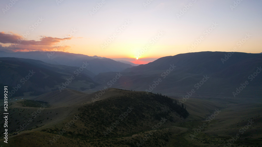 Red rays of the sun at sunset among the mountains. Green grass turns yellow, in places a small forest. Tourists walk in the hills. A river runs in the distance. The clouds are highlighted in pink.