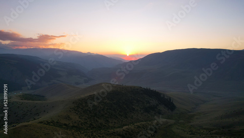 Red rays of the sun at sunset among the mountains. Green grass turns yellow  in places a small forest. Tourists walk in the hills. A river runs in the distance. The clouds are highlighted in pink.