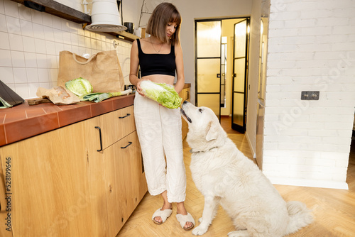 Young woman spends leisure time with her white dog while cooking healthy food in kitchen at modern apartment. Healthy eating and friendship with pets concept