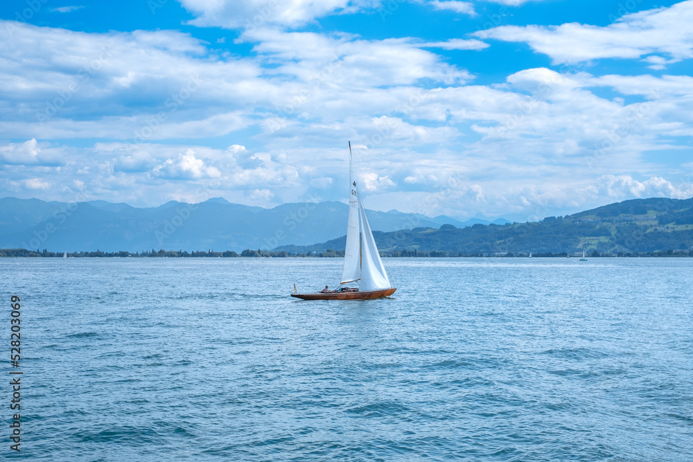 A small sailboat in a sea (Bodensee, Switzerland) with a blue sky with clouds and land in the background.