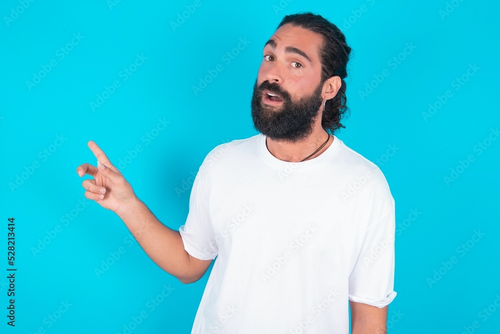 Emotive young bearded man wearing white T-shirt over blue studio wall keeps jaw dropped from shock demonstrates amazing promo points right on blank space demonstrates big shopping sale. Advertisement Emotive young bearded man wearing white T-shirt over blue studio wall keeps jaw dropped from shock demonstrates amazing promo points right on blank space demonstrates big shopping sale. Advertisement