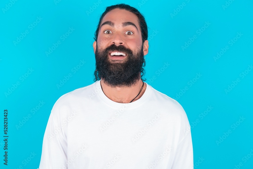 Shocked young bearded man wearing white T-shirt over blue studio background stares bugged eyes keeps mouth opened has surprised expression. Omg concept Shocked young bearded man wearing white T-shirt over blue studio background stares bugged eyes keeps mouth opened has surprised expression. Omg concept