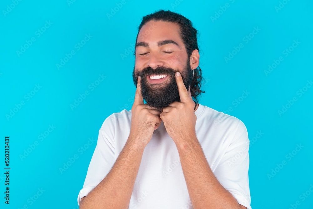 Happy young bearded man wearing white T-shirt over blue studio background with toothy smile, keeps index fingers near mouth, fingers pointing and forcing cheerful smile Happy young bearded man wearing white T-shirt over blue studio background with toothy smile, keeps index fingers near mouth, fingers pointing and forcing cheerful smile