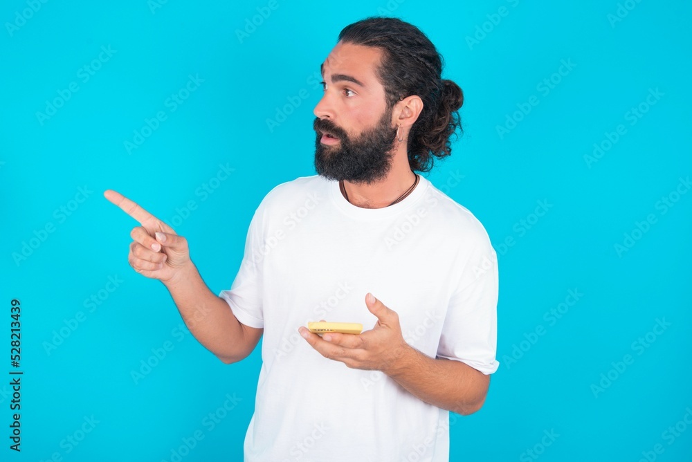 Fototapeta premium Stunned young bearded man wearing white T-shirt over blue studio background points sideways right copy space, recommends product, sees astonishing thing