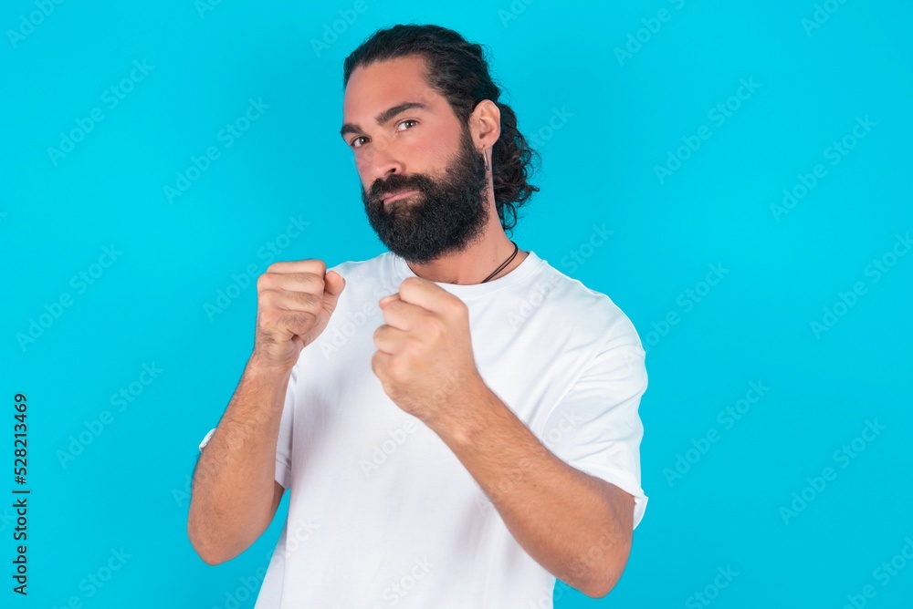 Displeased annoyed young bearded man wearing white T-shirt over blue studio wall clenches fists, gestures pissed, ready to revenge, looks with aggression at camera stands full of hate, being pressured Displeased annoyed young bearded man wearing white T-shirt over blue studio wall clenches fists, gestures pissed, ready to revenge, looks with aggression at camera stands full of hate, being pressured