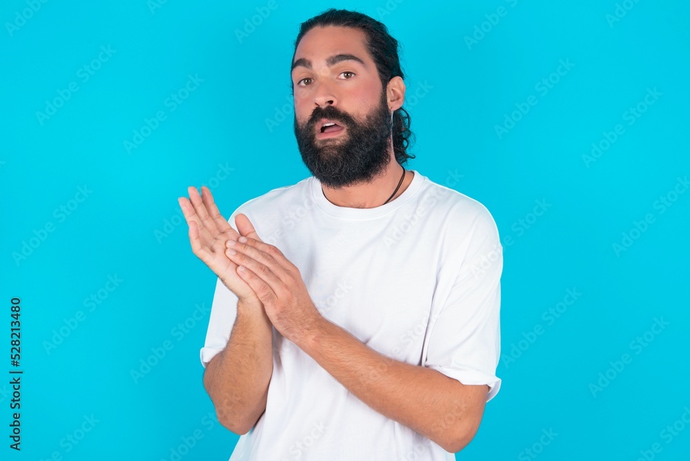 Surprised emotional young bearded man wearing white T-shirt over blue studio background rubs palms and stares at camera with disbelief Surprised emotional young bearded man wearing white T-shirt over blue studio background rubs palms and stares at camera with disbelief