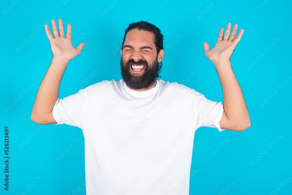 young bearded man wearing white T-shirt over blue studio background goes crazy as head goes around feels stressed because of horrible situation young bearded man wearing white T-shirt over blue studio background goes crazy as head goes around feels stressed because of horrible situation
