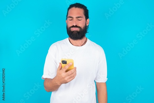 Upset dissatisfied young bearded man wearing white T-shirt over blue studio background uses mobile software application and surfs information in internet, holds modern mobile hand © Roquillo