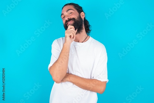 young bearded man wearing white T-shirt over blue studio background looking confident at the camera smiling with crossed arms and hand raised on chin. Thinking positive.
