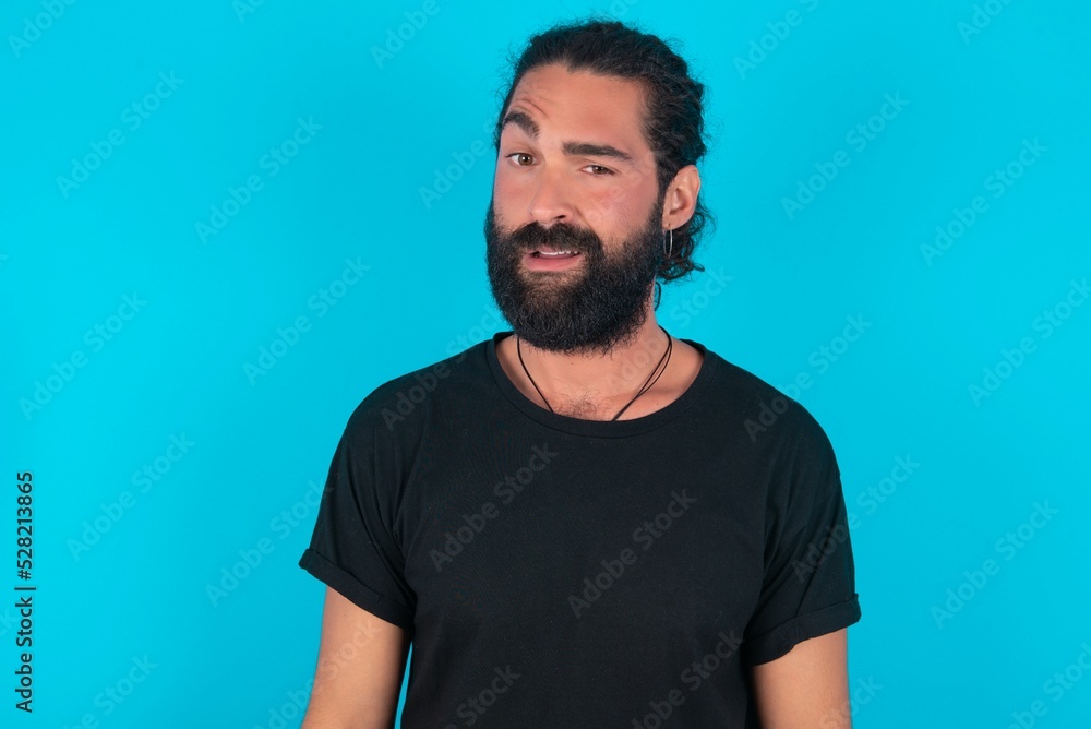 Portrait of dissatisfied young bearded man wearing black T-shirt over blue studio background smirks face, purses lips and looks with annoyance at camera, discontent hearing something unpleasant Portrait of dissatisfied young bearded man wearing black T-shirt over blue studio background smirks face, purses lips and looks with annoyance at camera, discontent hearing something unpleasant