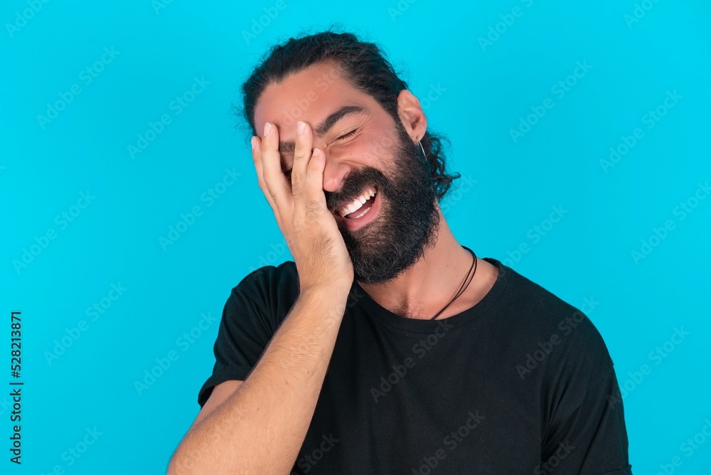 young bearded man wearing black T-shirt over blue studio background makes face palm and smiles broadly, giggles positively hears funny joke poses young bearded man wearing black T-shirt over blue studio background makes face palm and smiles broadly, giggles positively hears funny joke poses