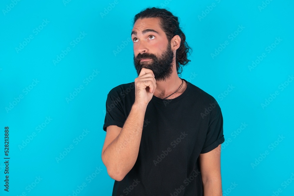 Thoughtful young bearded man wearing black T-shirt over blue studio background holds chin and looks away pensively makes up great plan Thoughtful young bearded man wearing black T-shirt over blue studio background holds chin and looks away pensively makes up great plan