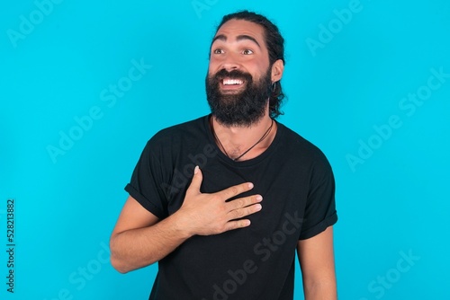 young bearded man wearing black T-shirt over blue studio background smiles toothily cannot believe eyes expresses good emotions and surprisement photo