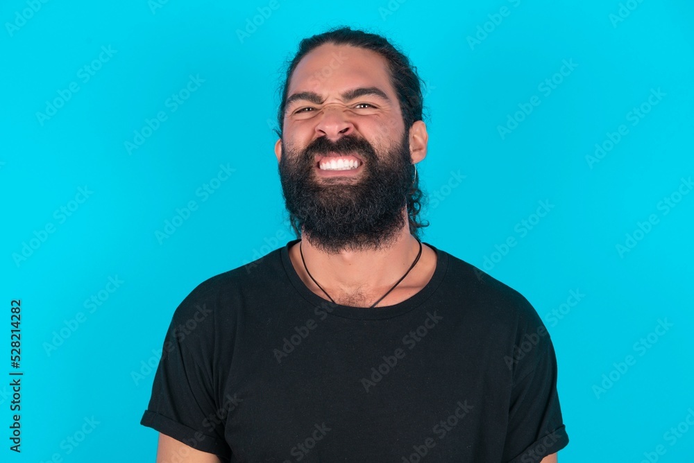Obraz premium young bearded man wearing black T-shirt over blue studio background keeps teeth clenched, frowns face in dissatisfaction, irritated because of much duties.