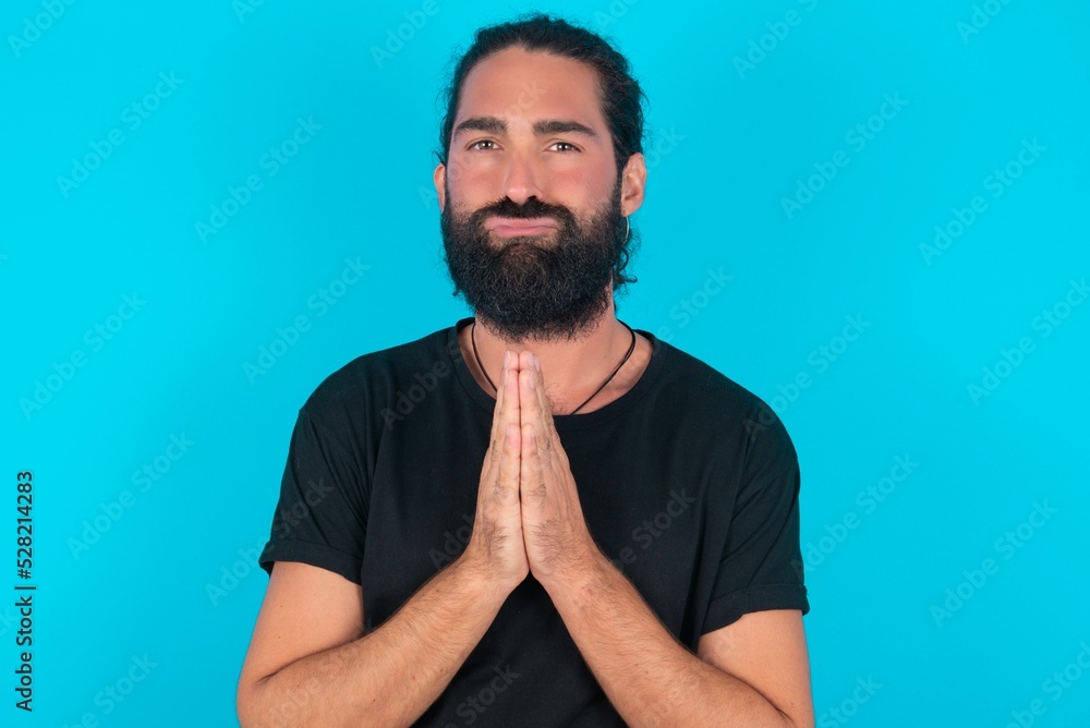 Positive young bearded man wearing black T-shirt over blue studio background smiles happily, glad to receive pleasant news from interlocutor, keeps palms together, People emotions concept. Positive young bearded man wearing black T-shirt over blue studio background smiles happily, glad to receive pleasant news from interlocutor, keeps palms together, People emotions concept.