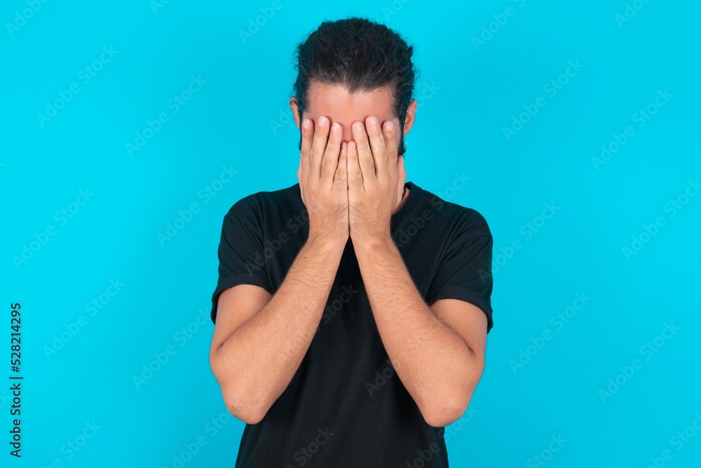 young bearded man wearing black T-shirt over blue studio background covering her face with her hands, being devastated and crying. Sad concept young bearded man wearing black T-shirt over blue studio background covering her face with her hands, being devastated and crying. Sad concept