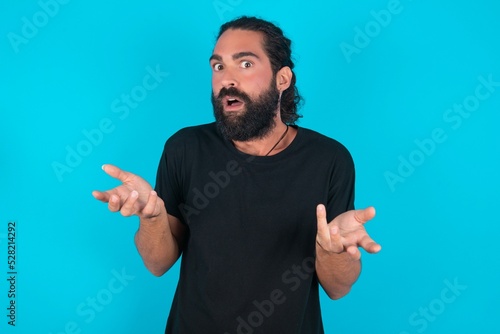 Frustrated young bearded man wearing black T-shirt over blue studio background feels puzzled and hesitant, shrugs shoulders in bewilderment, keeps mouth widely opened, doesn't know what to do.