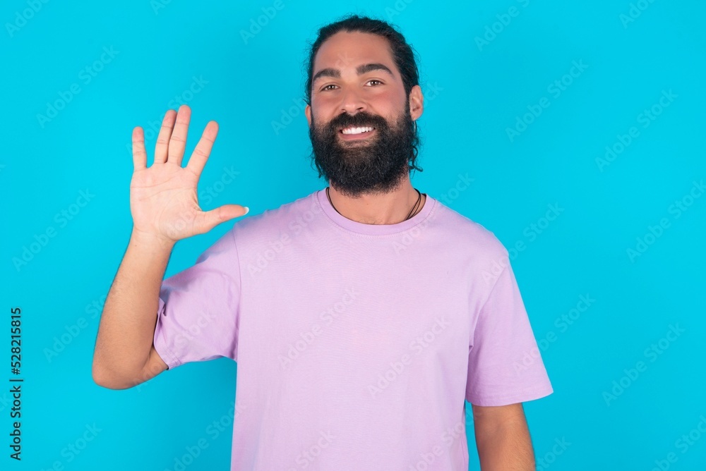 young bearded man wearing violet T-shirt over blue studio background showing and pointing up with fingers number five while smiling confident and happy. young bearded man wearing violet T-shirt over blue studio background showing and pointing up with fingers number five while smiling confident and happy.