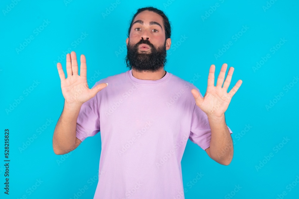 Naklejka premium young bearded man wearing violet T-shirt over blue studio background Moving away hands palms showing refusal and denial with afraid and disgusting expression. Stop and forbidden.