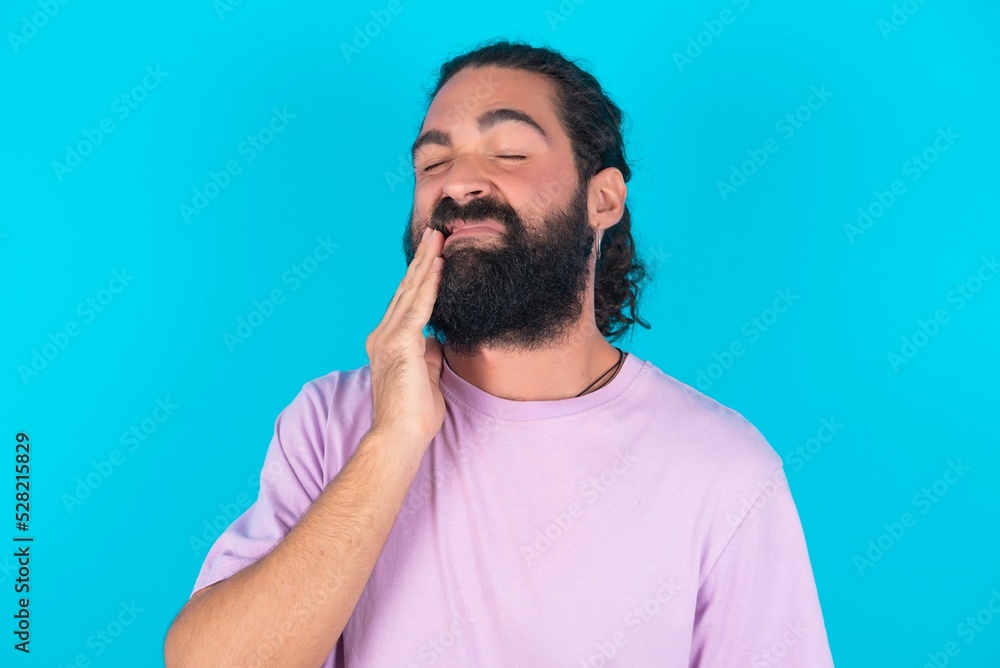 young bearded man wearing violet T-shirt over blue studio background touching mouth with hand with painful expression because of toothache or dental illness on teeth. young bearded man wearing violet T-shirt over blue studio background touching mouth with hand with painful expression because of toothache or dental illness on teeth.