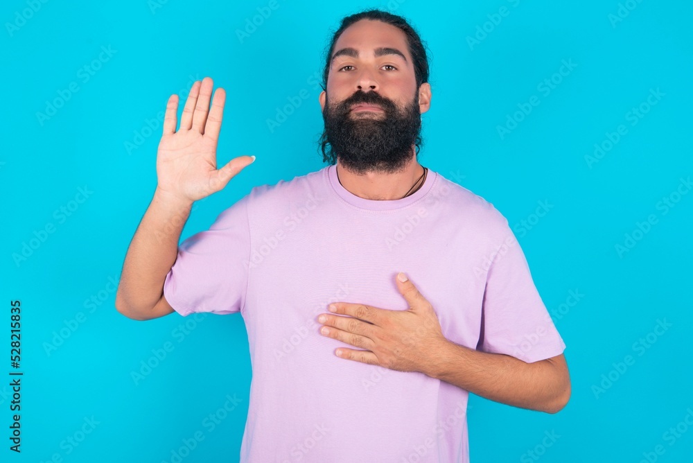 young bearded man wearing violet T-shirt over blue studio background Swearing with hand on chest and open palm, making a loyalty promise oath young bearded man wearing violet T-shirt over blue studio background Swearing with hand on chest and open palm, making a loyalty promise oath