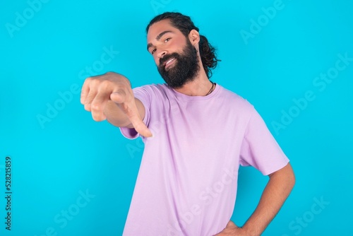 young bearded man wearing violet T-shirt over blue studio background pointing at camera with a satisfied, confident, friendly smile, choosing you