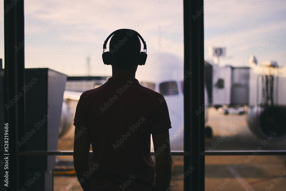 Fototapeta premium Silhouette of man with headphones while waiting for flight. Traveler looking from window of airport terminal before boarding to airplane..