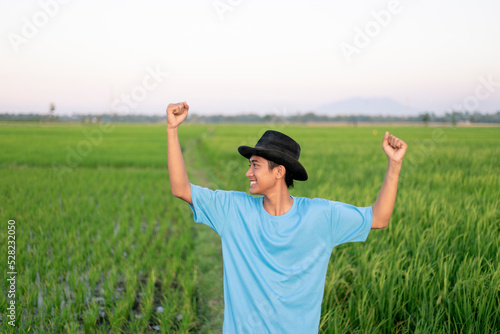 Attractive cheerful young Asian farmer raising arms rejoicing having success attainment to growing paddy rice at the rice field.