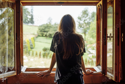 Rearview of long dark-haired woman in t shirt looking out the window, contemplating landscape rustic natural tree view #528233612