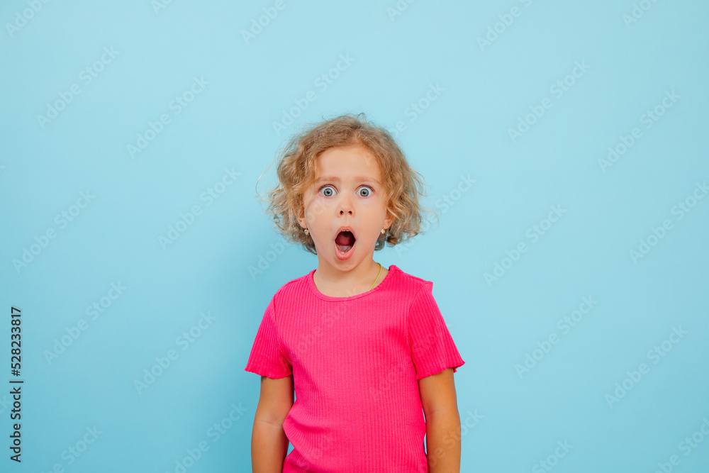 Little expressive, shocked, emotional curly blonde girl in pink shirt with open mouth, look at camera on blue view Little expressive, shocked, emotional curly blonde girl in pink shirt with open mouth, look at camera on blue view