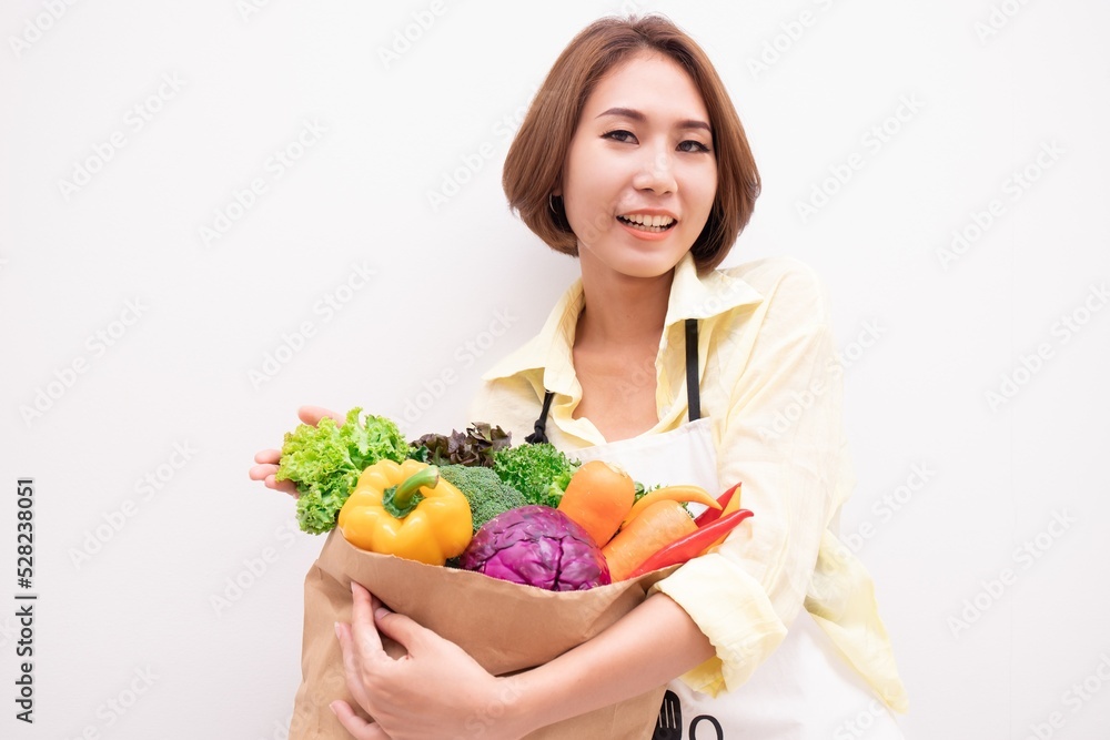 Young Asian woman holding grocery paper shopping bag full of fresh vegetables. Diet healthy eating concept isolated on a white background. Young Asian woman holding grocery paper shopping bag full of fresh vegetables. Diet healthy eating concept isolated on a white background.