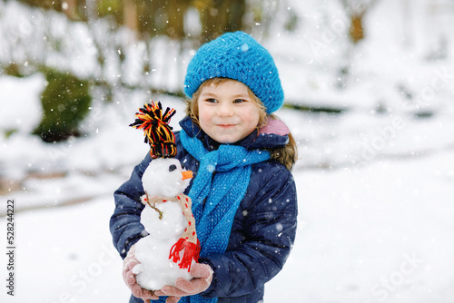Cute little toddler girl making mini snowman and eating carrot nose. Adorable healthy happy child playing and having fun with snow, outdoors on cold day. Active leisure with children in winter