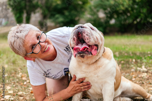 Senior woman sitting on ground and hugging english bulldog looking at camera outdoors. Woman playing with dog in park