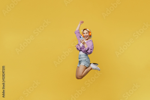 Full body side view young blonde woman 20s in nk tied shirt white t-shirt jump high hold use mobile cell phone do winner gesture isolated on plain yellow background studio People lifestyle concept © ViDi Studio