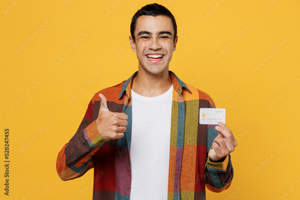 Young smiling happy middle eastern man 20s wear casual shirt white t-shirt hold in hand mock up of credit bank card show thumb up isolated on plain yellow background studio People lifestyle concept Young smiling happy middle eastern man 20s wear casual shirt white t-shirt hold in hand mock up of credit bank card show thumb up isolated on plain yellow background studio People lifestyle concept