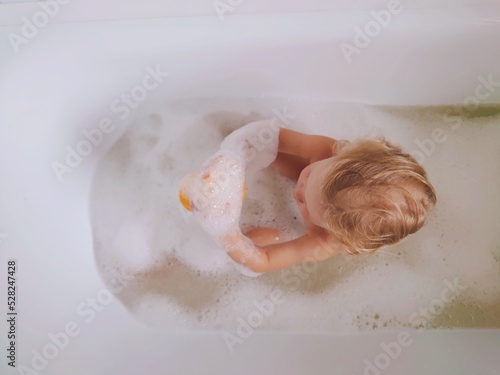 Child taking a babble bath. Cute and adorable toddler baby boy playing with foam and water. Natural light in a bathroom.
