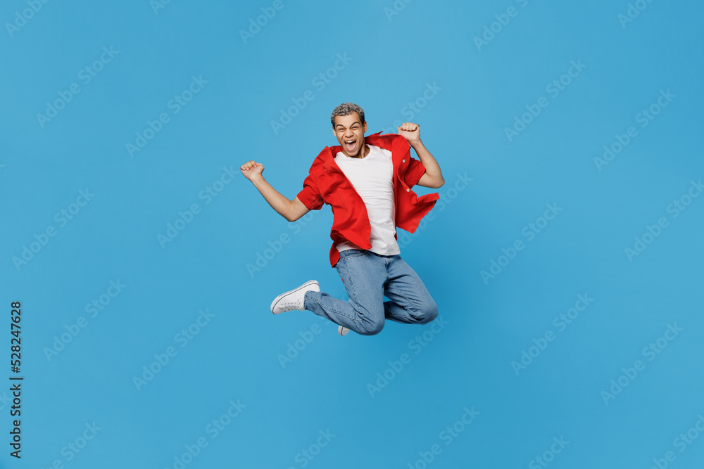 Full body young overjoyed excited cool man of African American ethnicity 20s wear red shirt jump high do winner gesture isolated on plain pastel light blue cyan background. People lifestyle concept