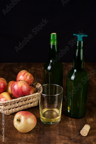 Bottles of cider, one with a stopper for pouring, next to a glass of cider and some apples, on a dark background and a wooden table.