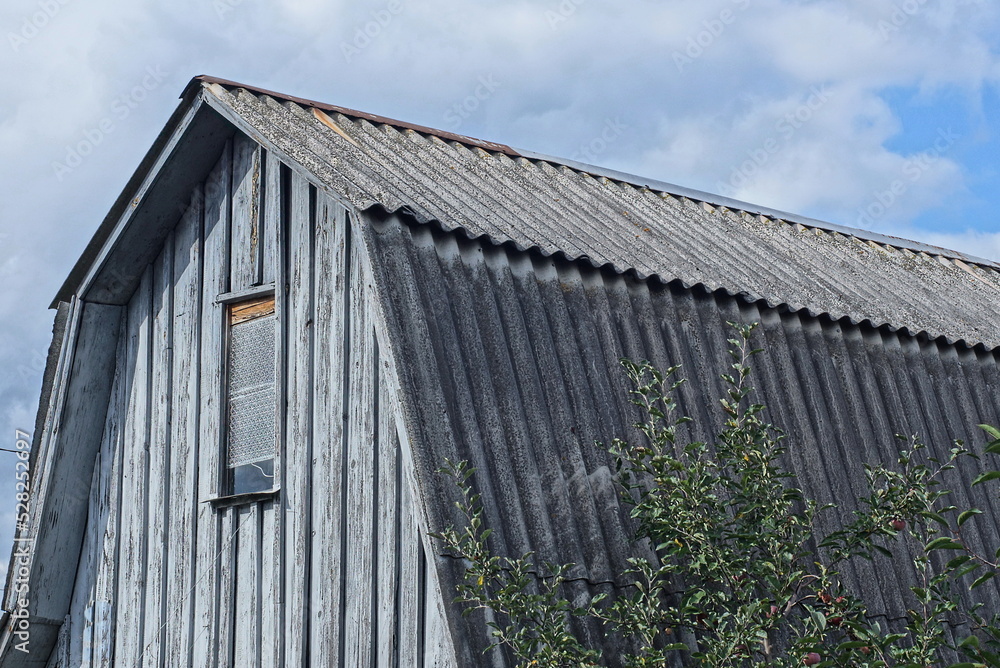 wooden attic of a rural house with a small window under a gray slate roof on the street against the sky