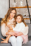 Mom and daughter on New Year's Eve are sitting in a Scandinavian-style room in light warm sweaters with long flowing hair on a gray sofa, happy and hugging