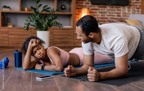 Young african american guy supports tired sad lady in sportswear doing plank exercises on mat