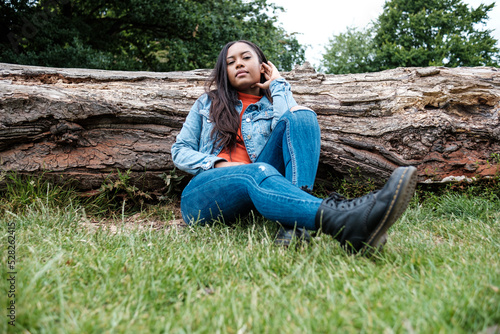 Portrait of young race mixed woman sitting on the grass and leaning against a fallen tree in a park.