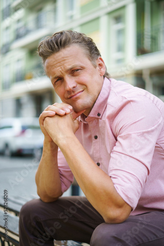 Handsome man spend time sitting on the bench in urban city with hands put together and leaned chin looking at camera, wearing pink shirt. Smiling freelancer caucasian man off work