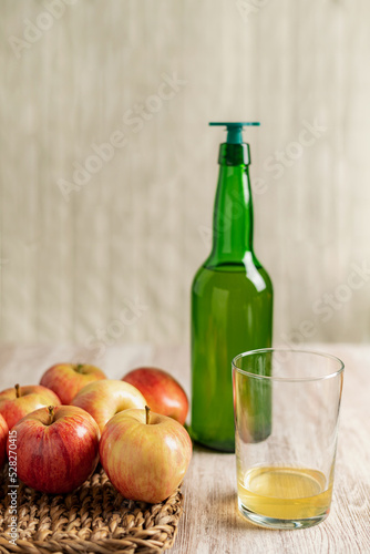 Glass with a culin of cider, next to a bottle with a pourer and some apples, on a light wood table.