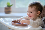 Happy toddler baby eats fruits and berries sitting on a high children chair. A child at the window with a beige plate of food. Kid aged one year and two months