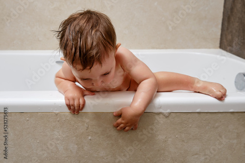 Toddler baby climbs over the edge of the tub at the risk of falling. The danger of leaving a child alone in the bathroom